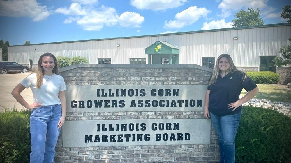 Two women are standing in front of a sign that says illinois corn growers association.