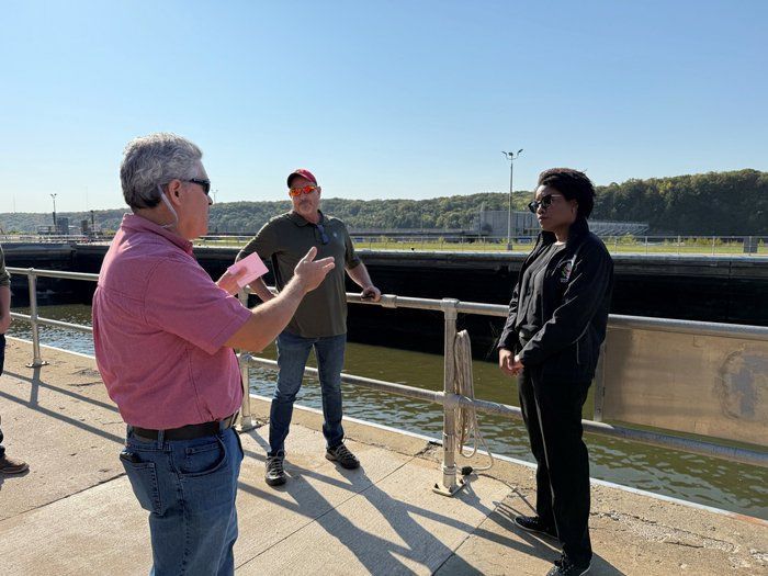 A man in a pink shirt is talking to a group of people standing next to a body of water.