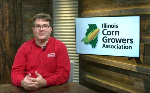 A man in a red shirt is sitting at a table in front of an illinois corn growers association sign.
