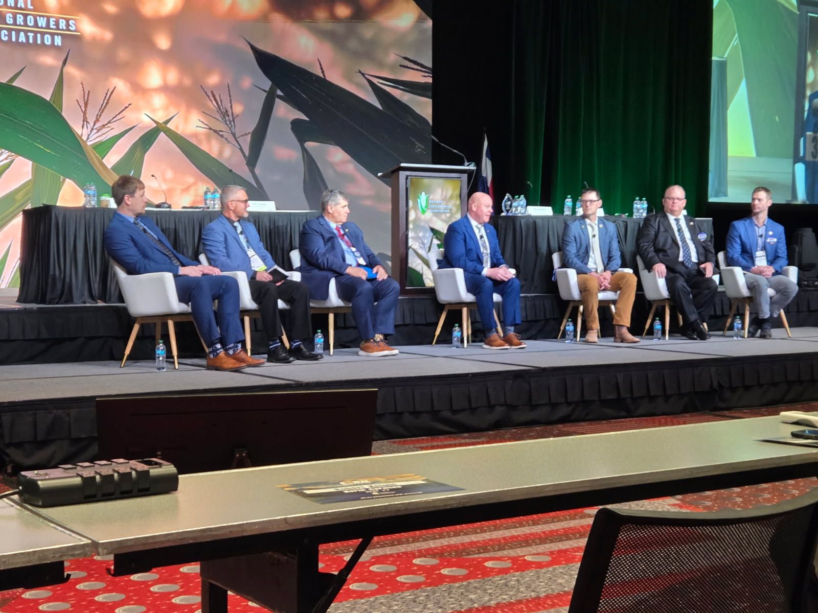 Panel of men in suits on stage; event backdrop with corn.