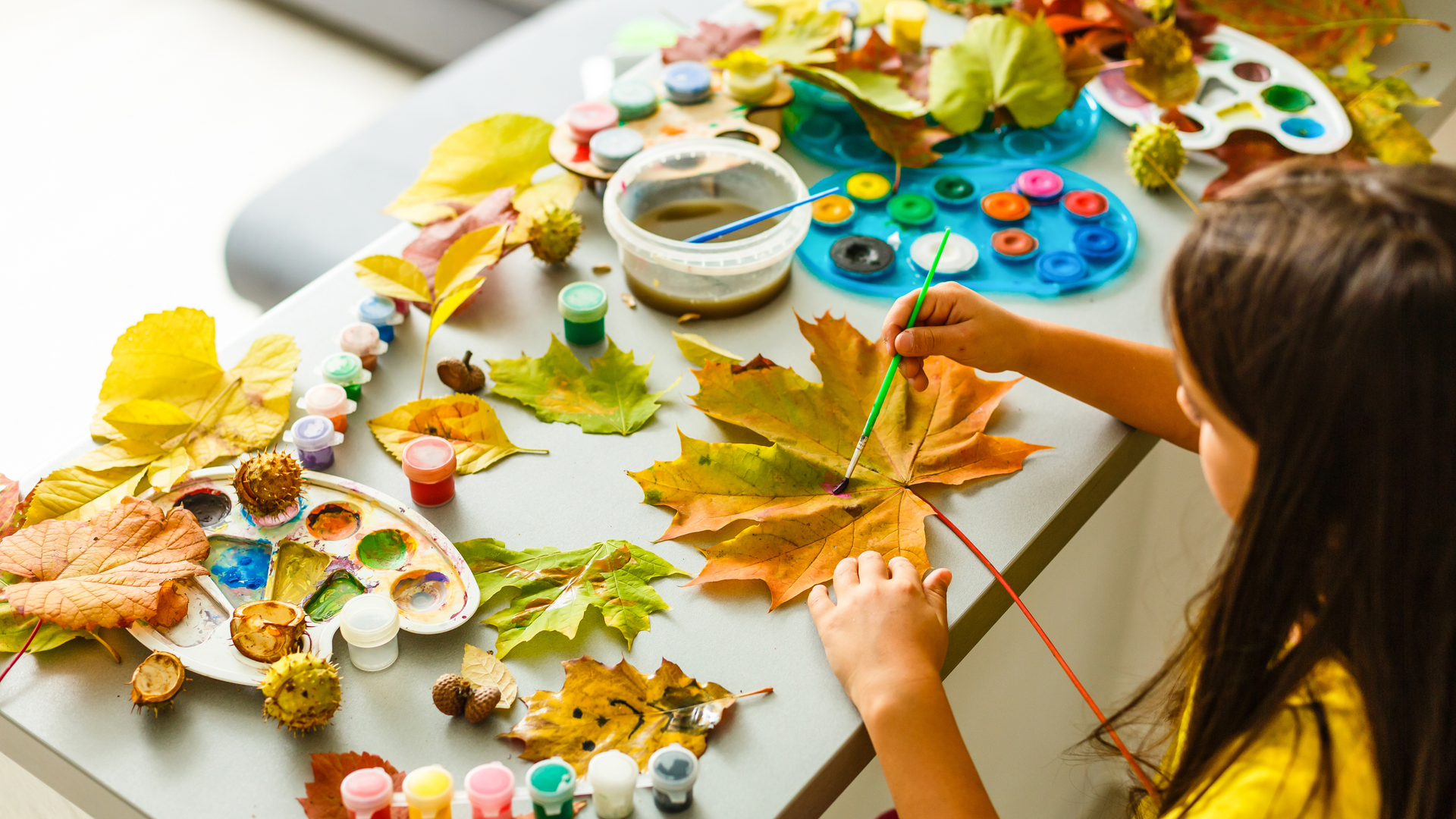 Girl painting a leaf with brush at a table with paints, leaves, and other art supplies.