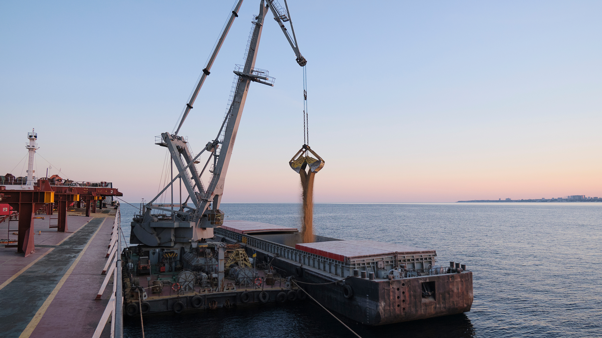 A crane loads grain from a dock onto a ship at a port, with a sunset sky over the sea.