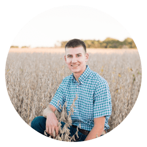 A young man in a plaid shirt is kneeling in a field of wheat.