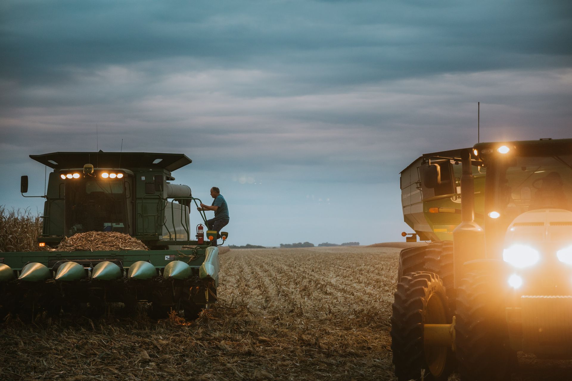 Harvesting equipment in a field at dusk. A farmer looks over the combine while lights shine.