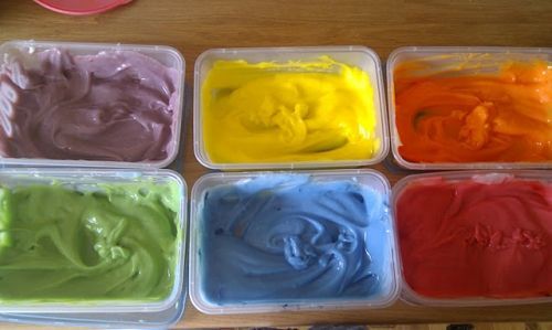 Six containers of different colored frosting are sitting on a wooden table.