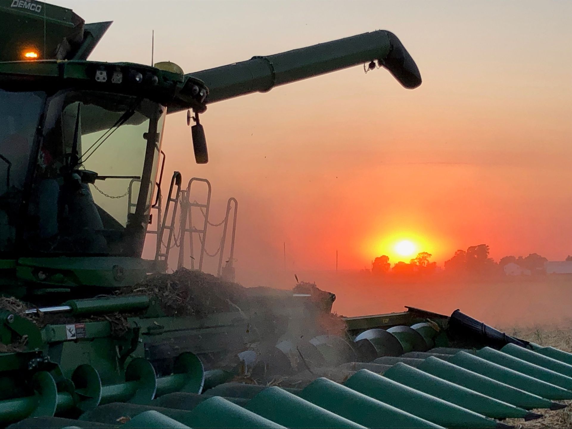 Combine harvester at sunset, harvesting crops, with orange and yellow sky.