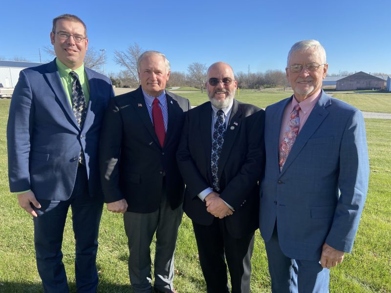Four men in suits and ties are posing for a picture in a grassy field.