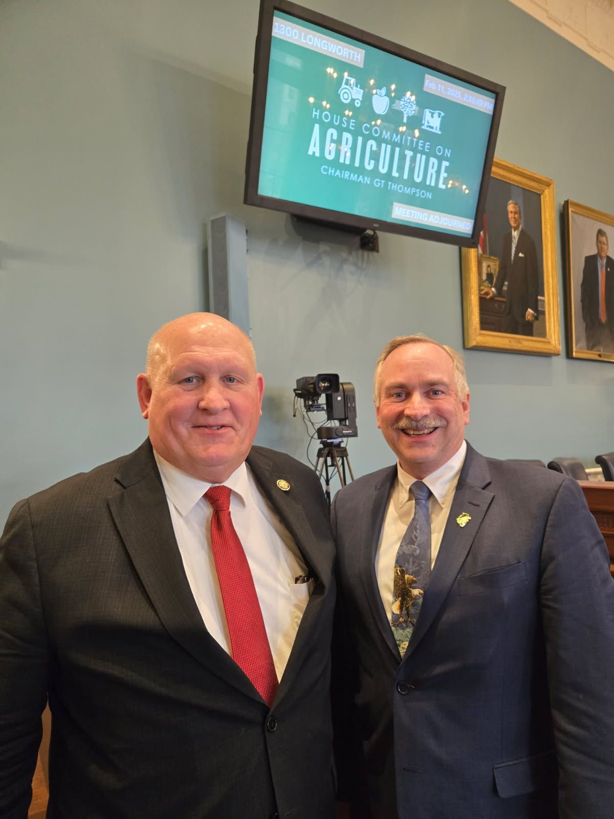 Two men in suits stand in a room, beneath a screen displaying