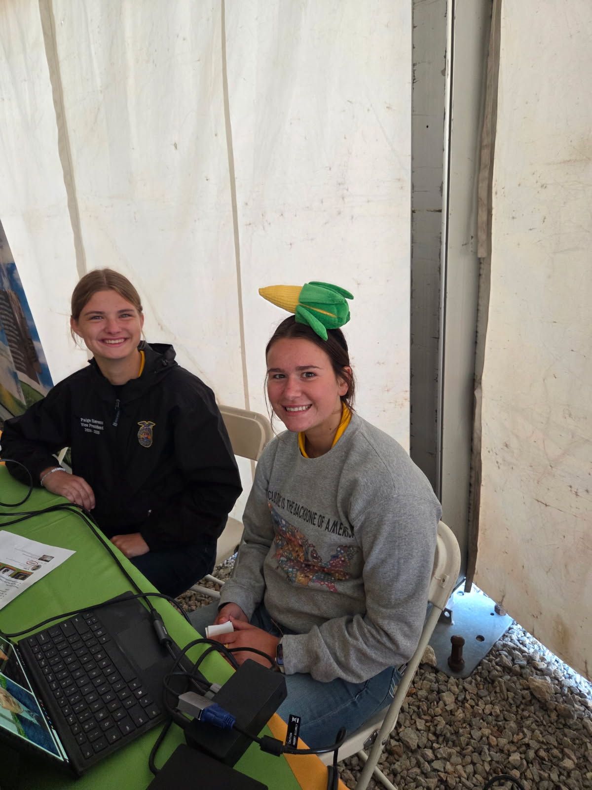 Two smiling women at a table; one wears a corn-on-the-cob hat. They sit inside a tent.