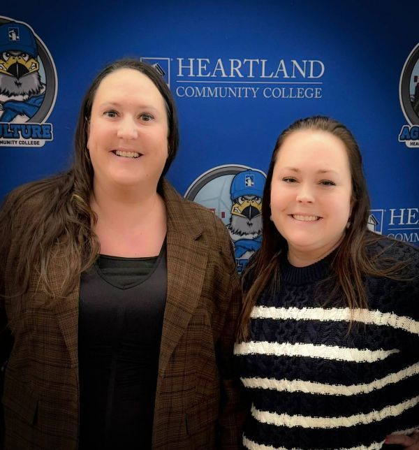 Two women are posing for a picture in front of a heartland community college banner