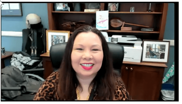 A woman is smiling while sitting in a chair in front of a shelf.