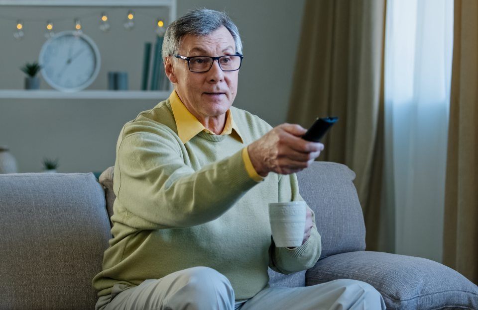 An elderly man is sitting on a couch holding a cup of coffee and a remote control.