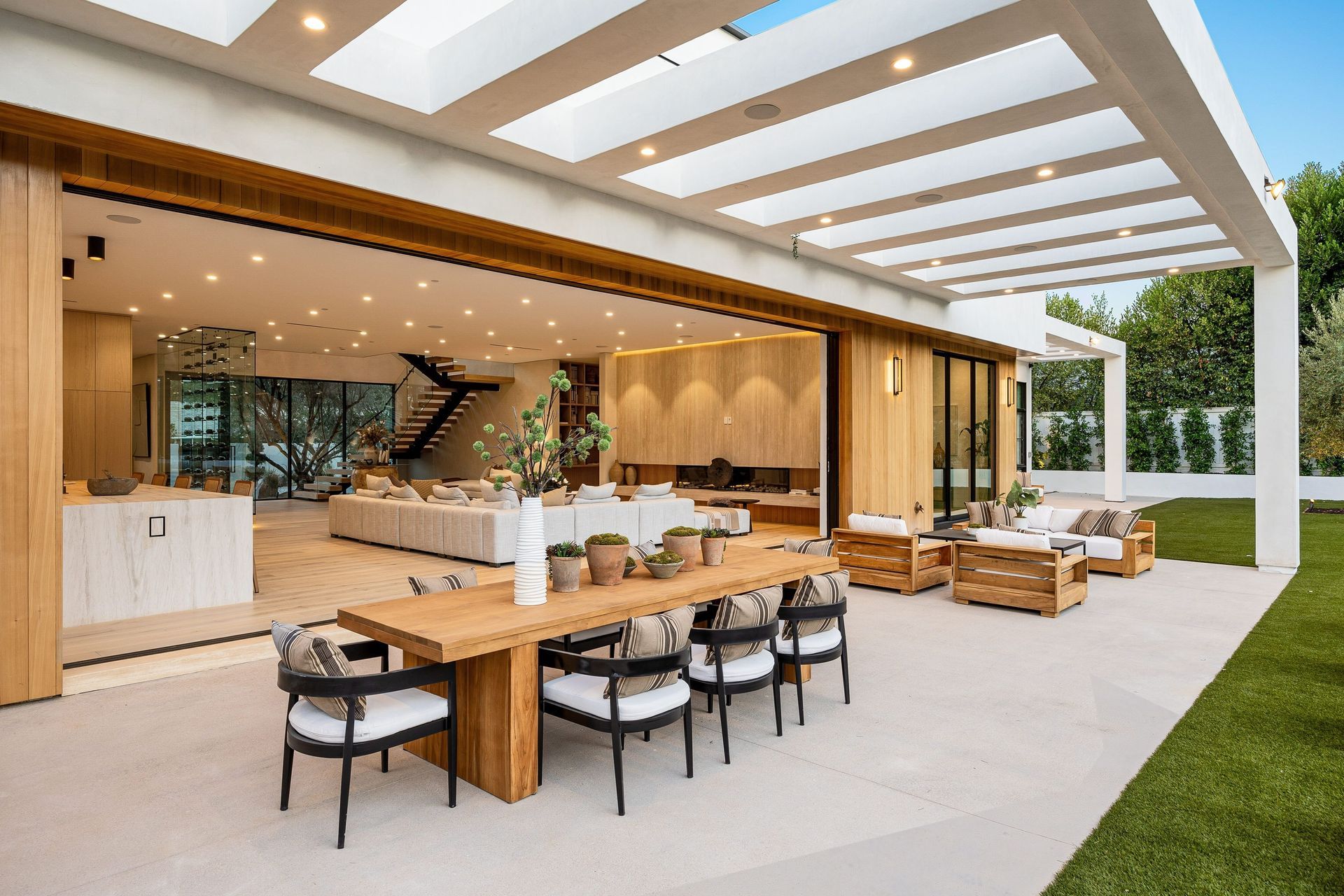 Outdoor patio with wooden dining table, lounge seating, and modern open-air kitchen under a slatted roof