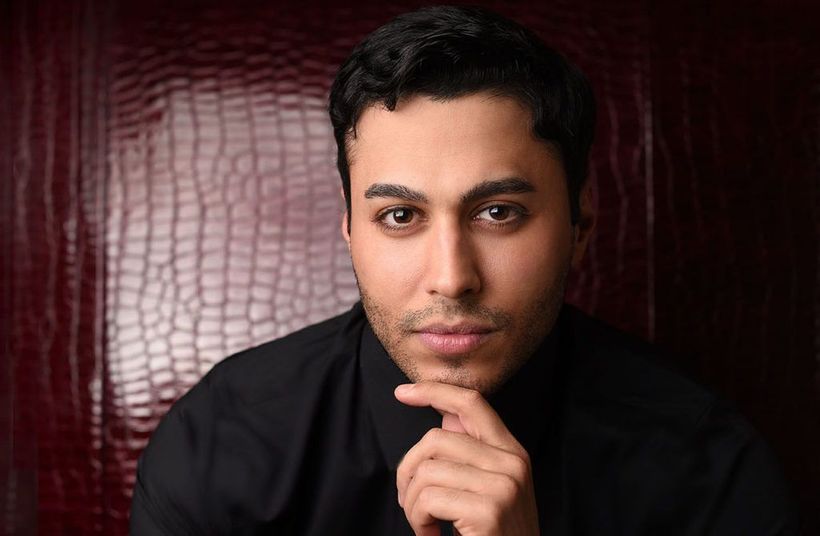 Man with dark hair and a black shirt, resting his chin on his hand, looking toward the camera.