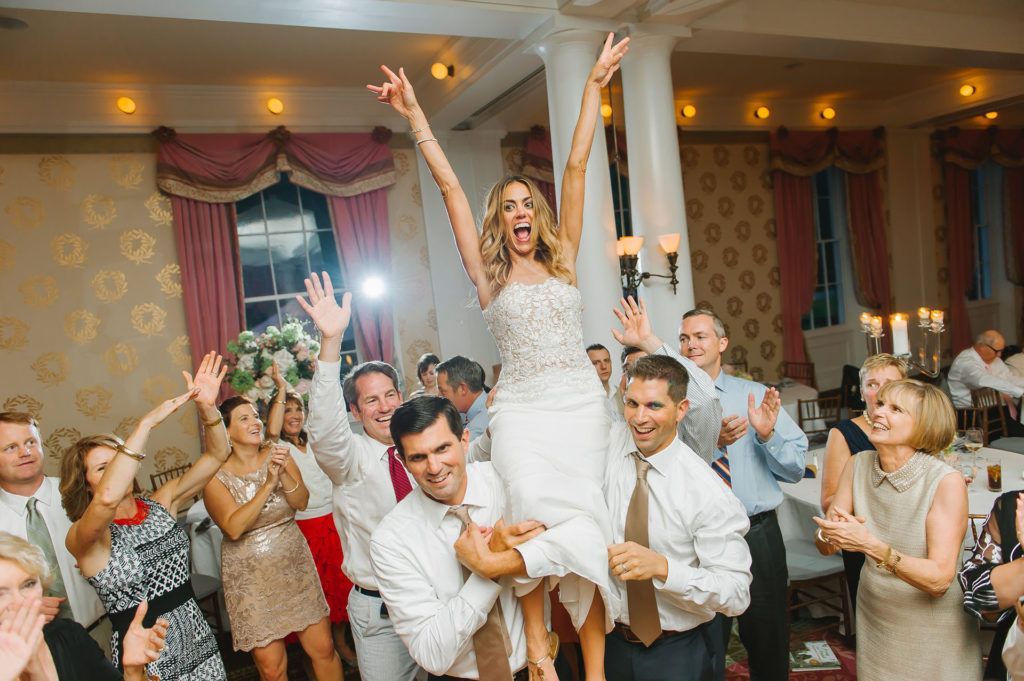 Bride is lifted on shoulders, arms raised, celebrating at a wedding reception. Other guests cheer in a ballroom setting.