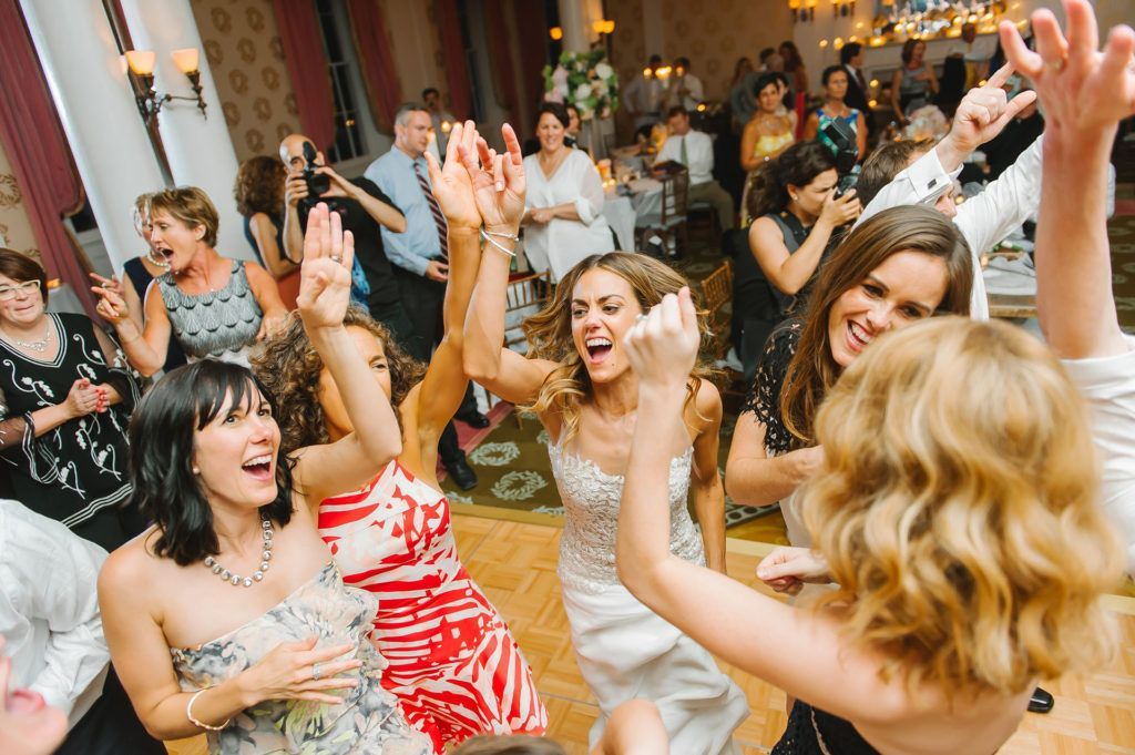 Wedding guests dancing enthusiastically on a wooden dance floor. People are raising their arms, smiling.