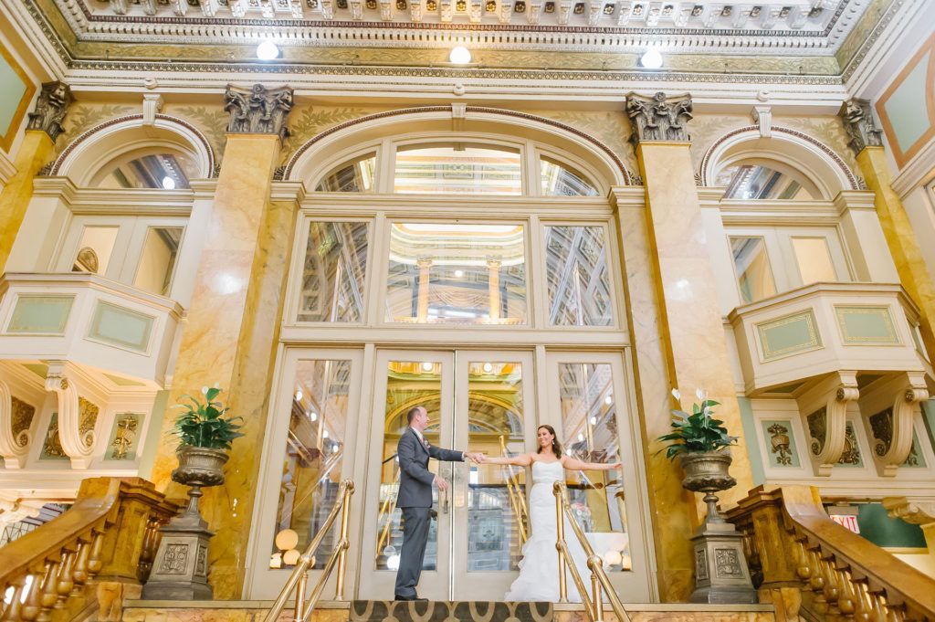 Bride and groom holding hands in ornate building doorway. They're standing on steps, facing the camera, with marble columns.