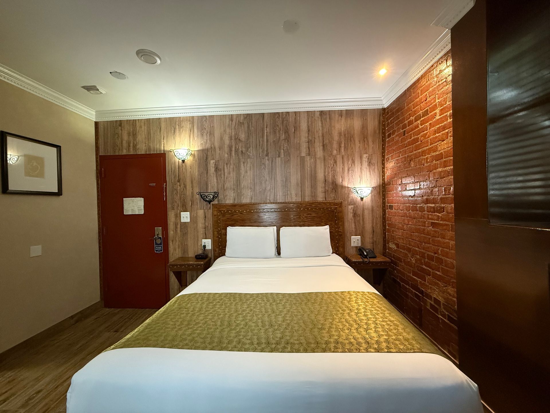 Bedroom with wood-paneled wall behind bed, brick wall on right, red door on left, bed with gold bed runner.