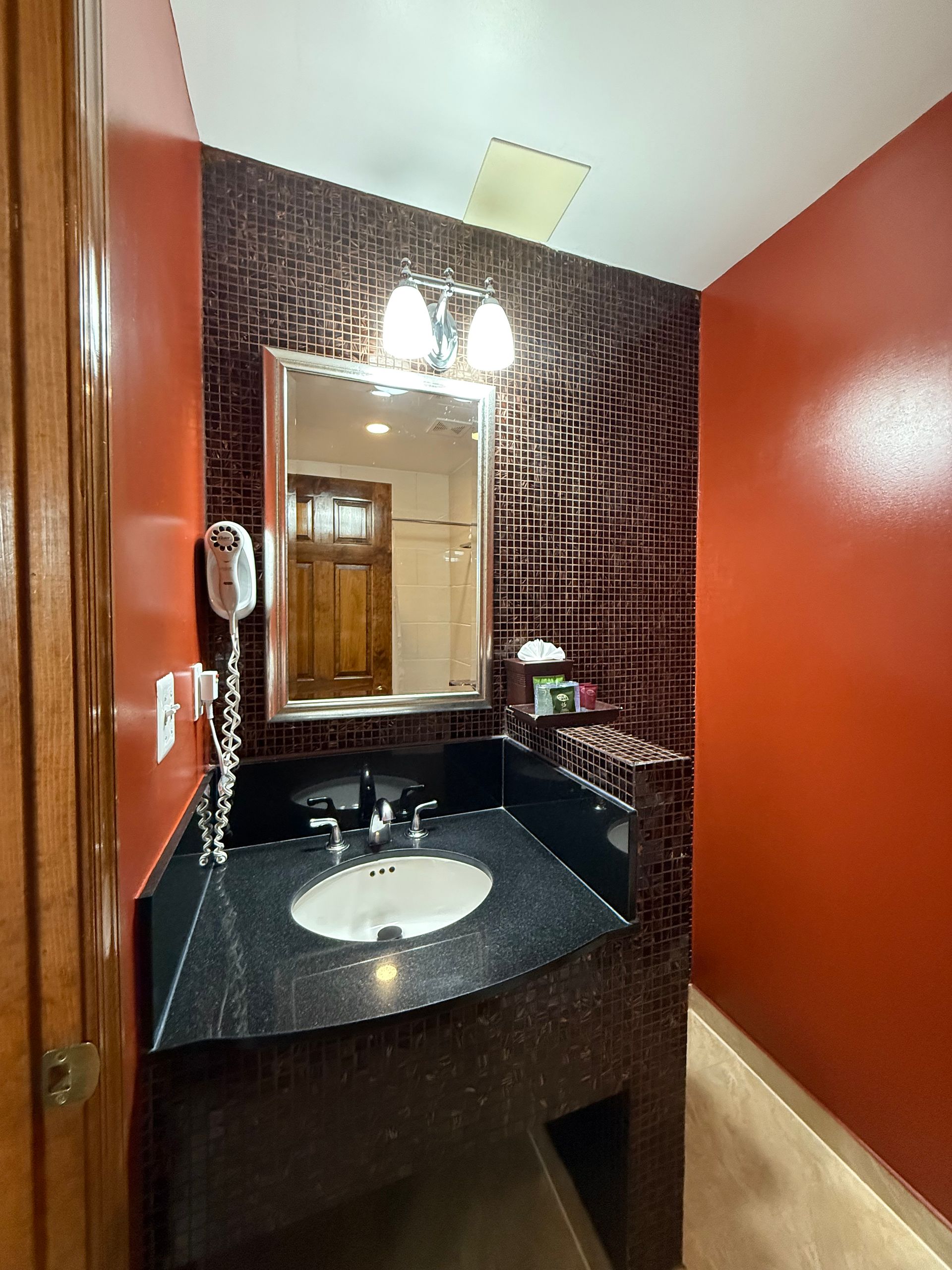Small bathroom with red walls, mosaic backsplash, black countertop, and silver-framed mirror.