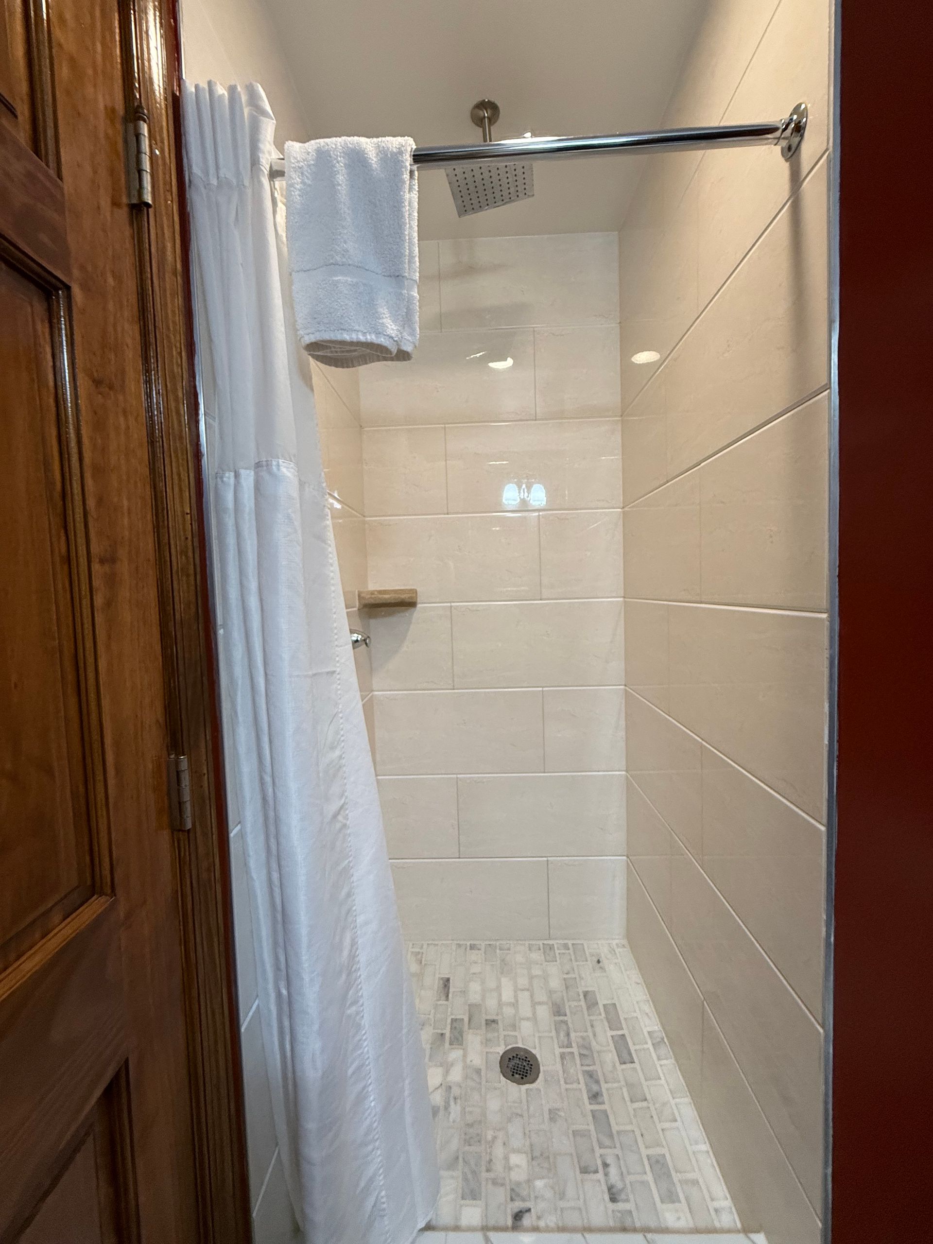 Shower stall with white tile, glass door, and white curtain; a wooden door is partially visible on the left.