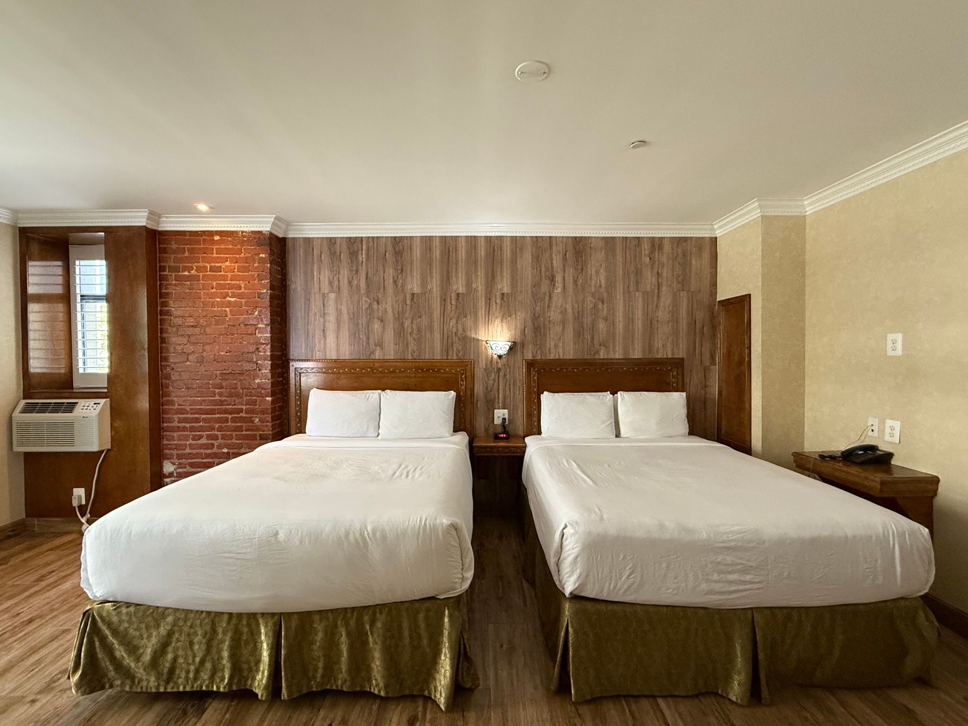 Two beds in a hotel room with wood paneling, brick, and gold bed skirts.