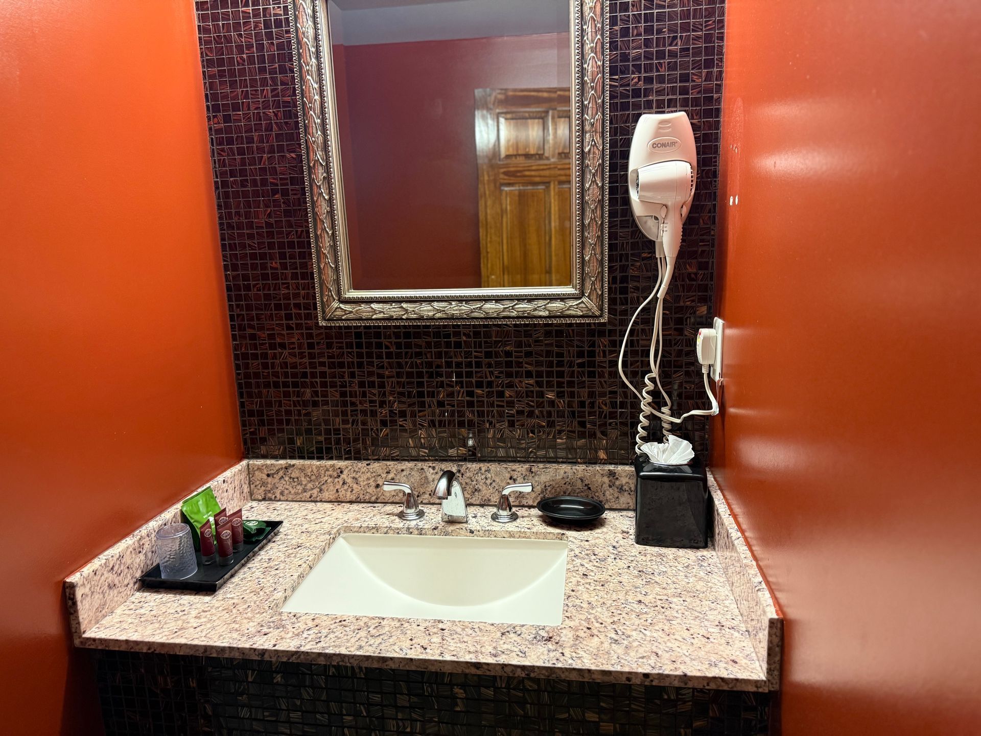 Bathroom with orange walls, mosaic backsplash, framed mirror, sink, and hairdryer.