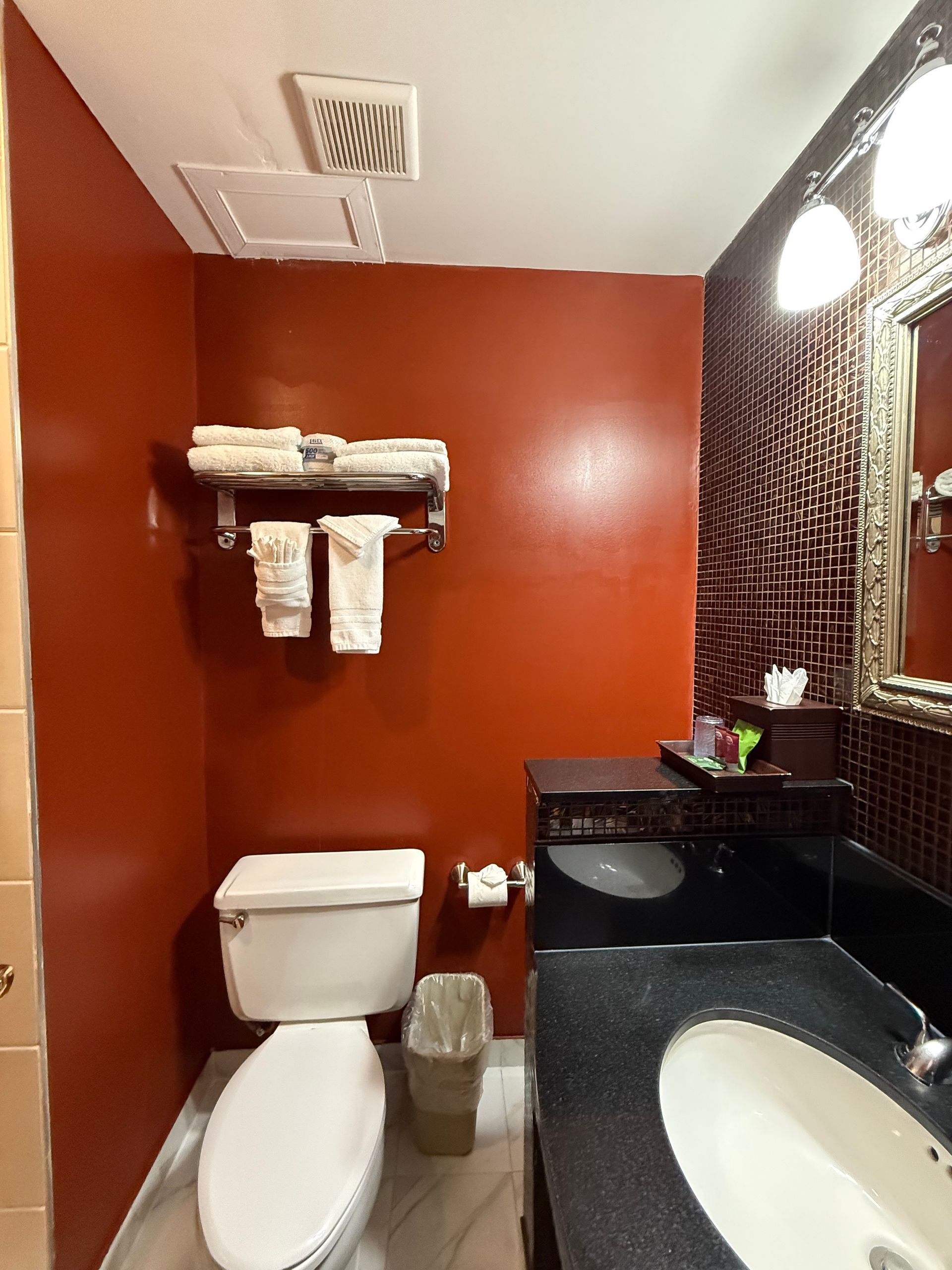Small bathroom with red walls, toilet, sink, towels, and a decorative mirror.