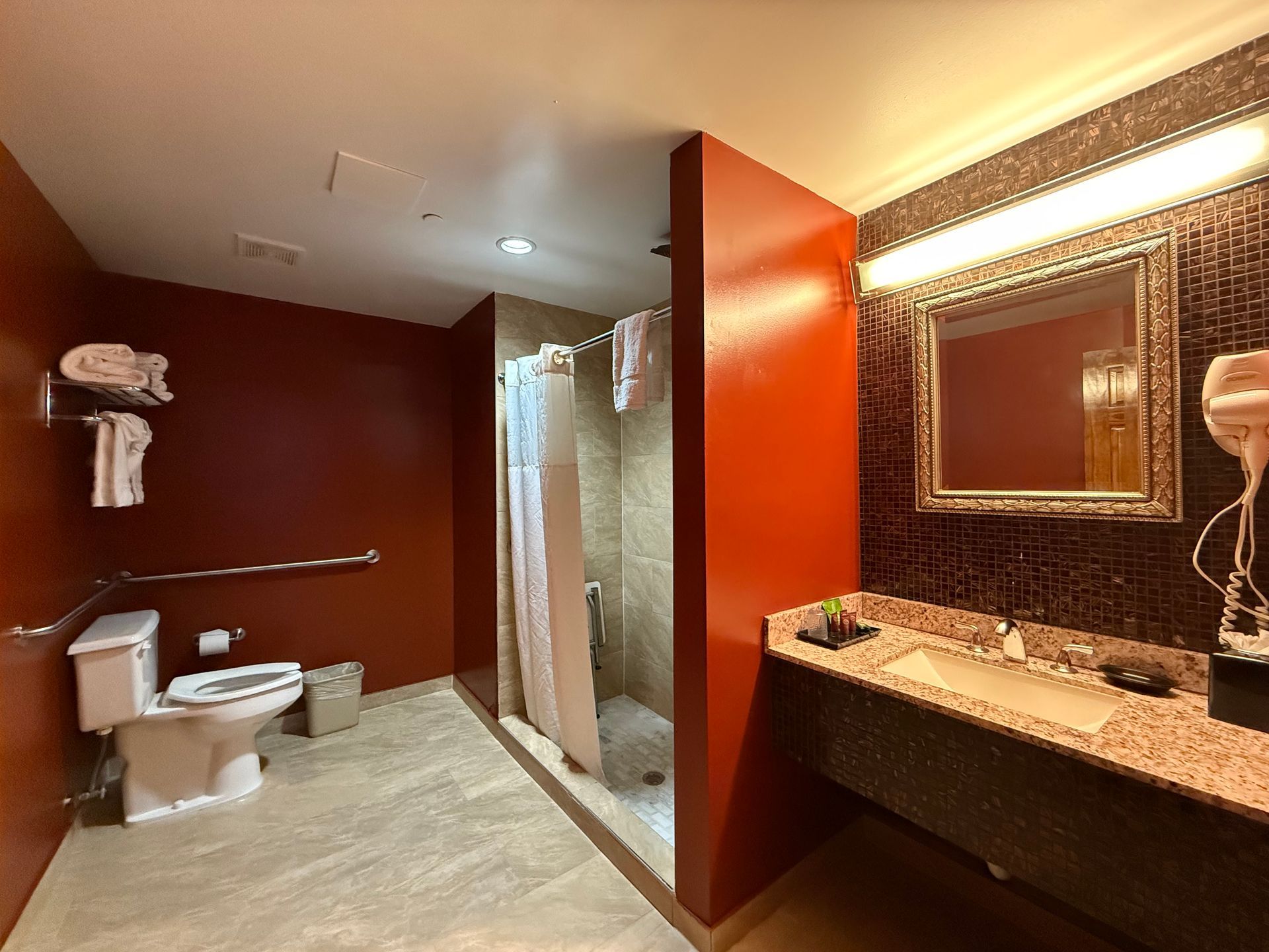 A hotel bathroom with red walls, a shower, sink, and toilet. Includes accessibility grab bar.
