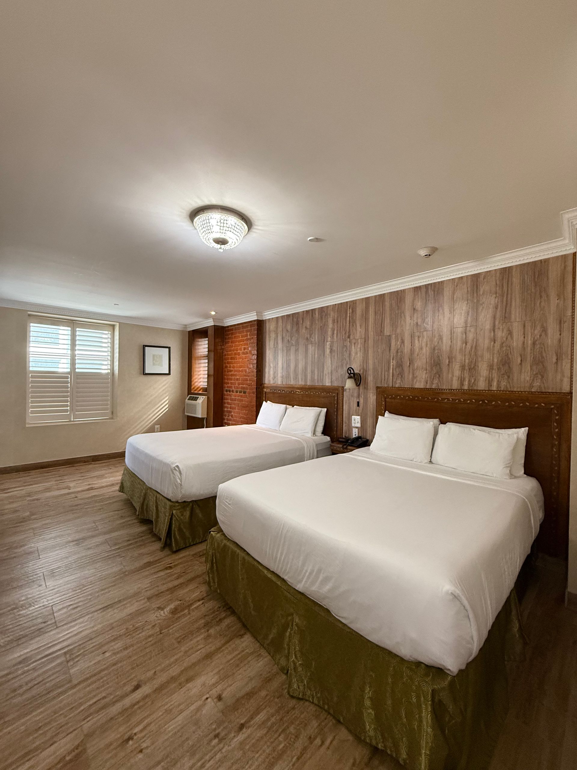 Two beds in a hotel room with wood-paneled wall, window, and light-colored flooring.
