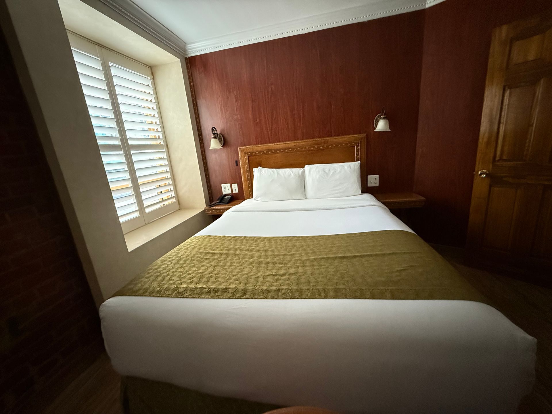 Bedroom with a queen-sized bed, shutters, and wooden door. Burgundy walls and a gold bed runner.