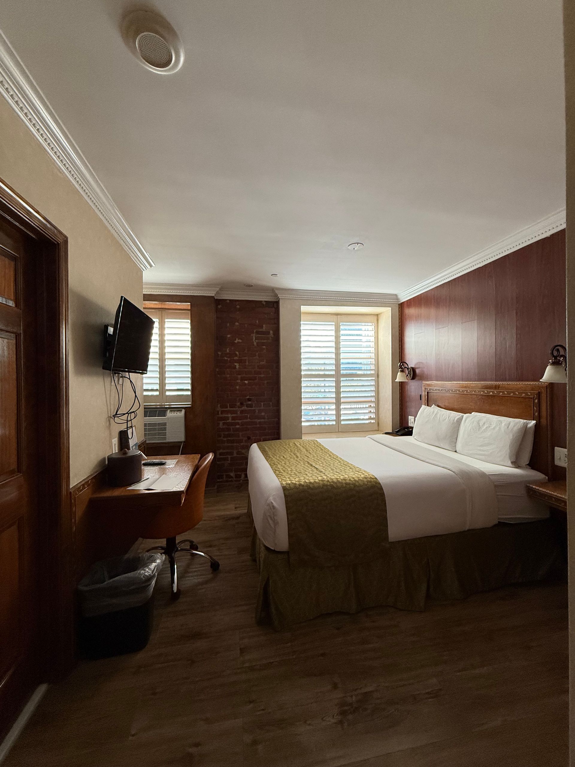 Hotel room with a bed, desk, TV, and two windows.