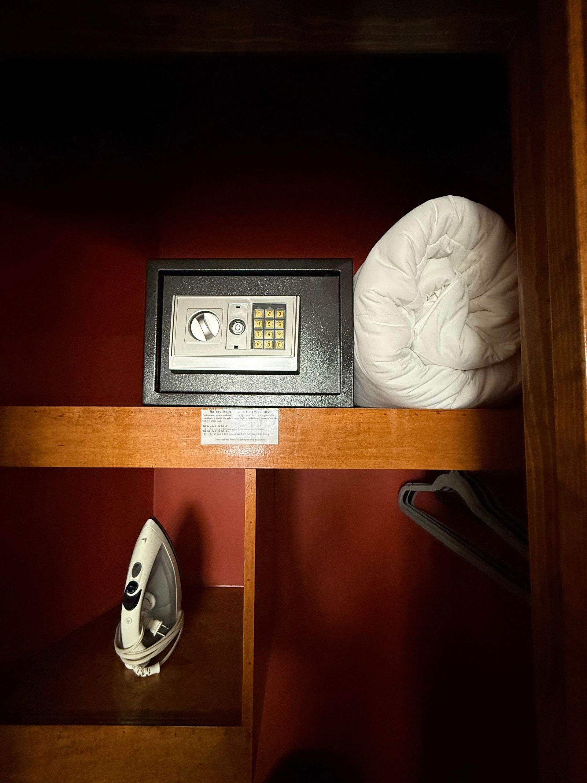 Interior of a closet with a safe, towels, an iron, and hangers on a shelf. Red walls, wood accents.