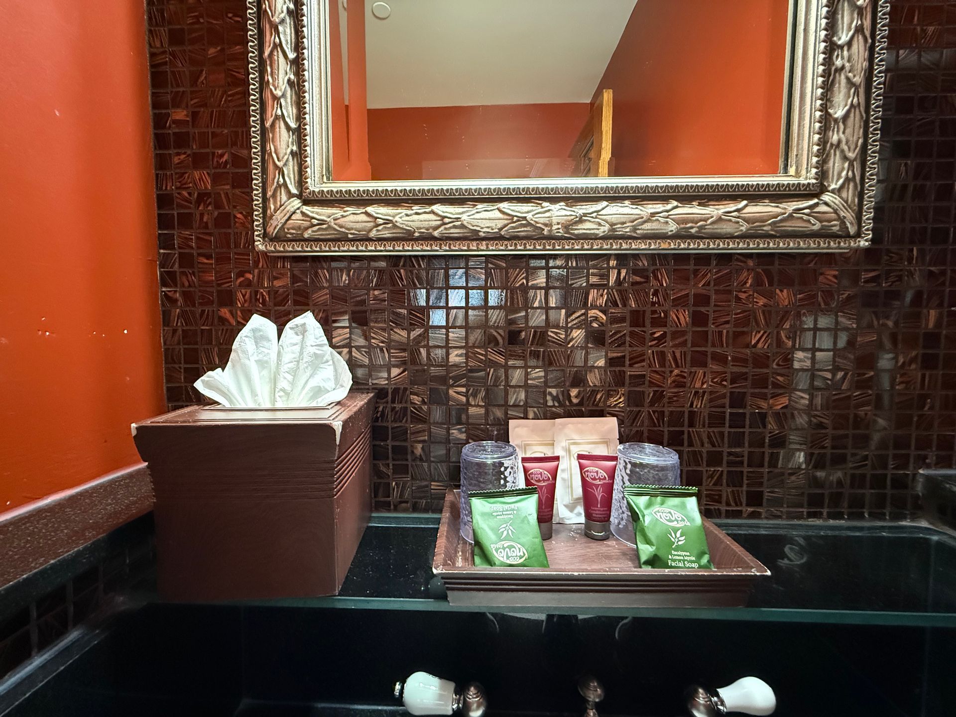 Bathroom vanity with mirror, tissue box, and toiletries on a tray, with a red-tiled backsplash.