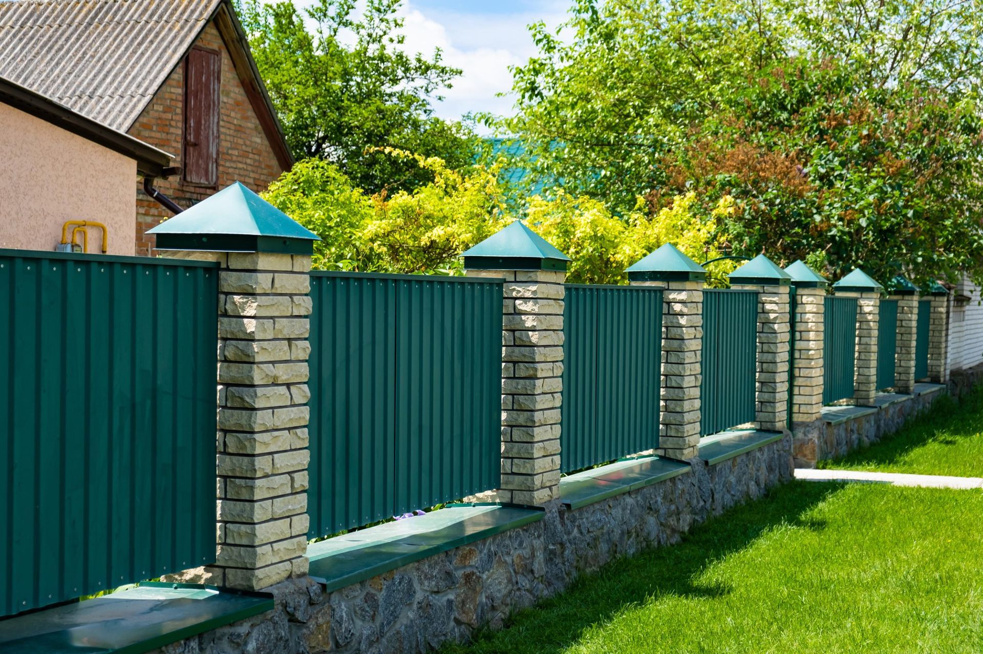 Green metal fence with brick pillars and decorative tops, in front of a house and trees.