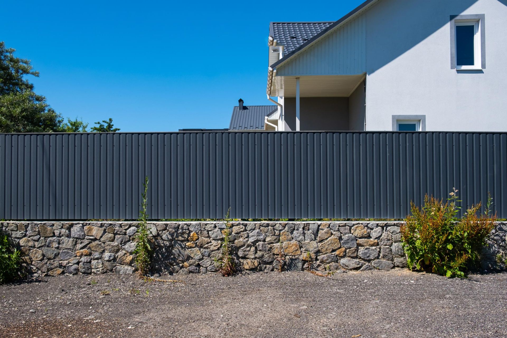 Gray metal fence atop a stone wall, in front of a light-colored house under a blue sky.