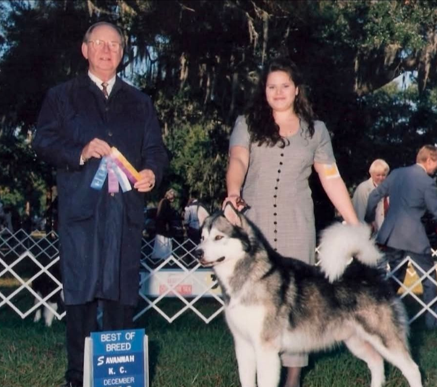 Two people posing with a husky at a dog show, with a blue award sign in front of them.