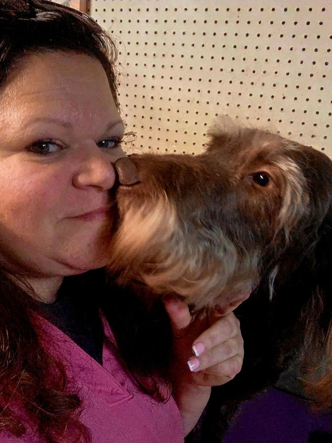 Woman in a pink top kissing a small brown dog indoors.