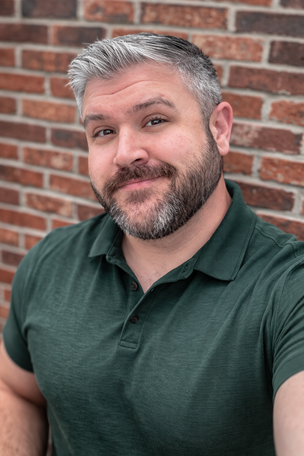 Man with glasses, graying hair, red and black plaid shirt, smiling, brick wall background.
