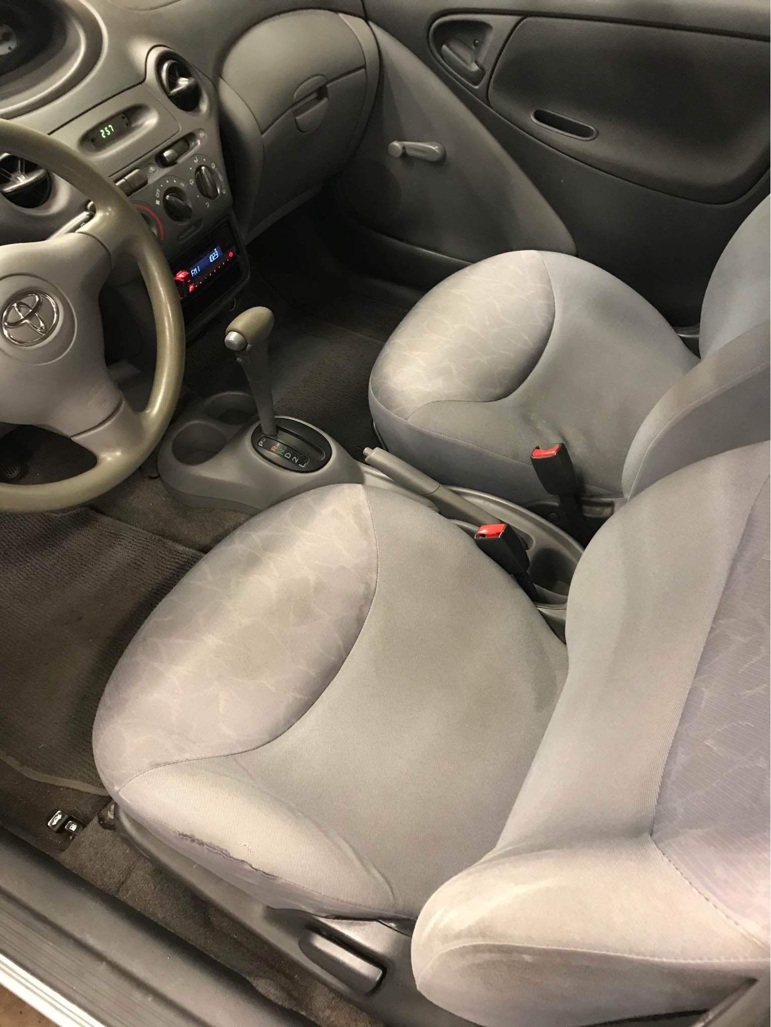 Interior of a Toyota Yaris: grey seats, dashboard, steering wheel. View of front seats and center console.