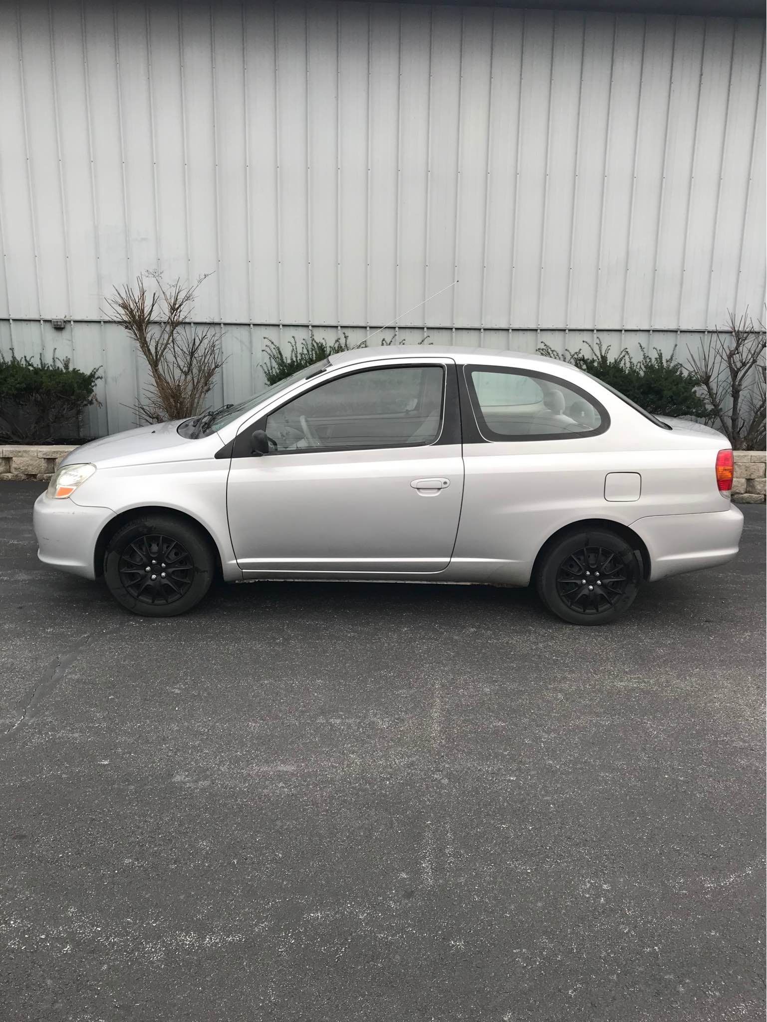 Silver Toyota Echo parked in front of a gray building, black wheels.