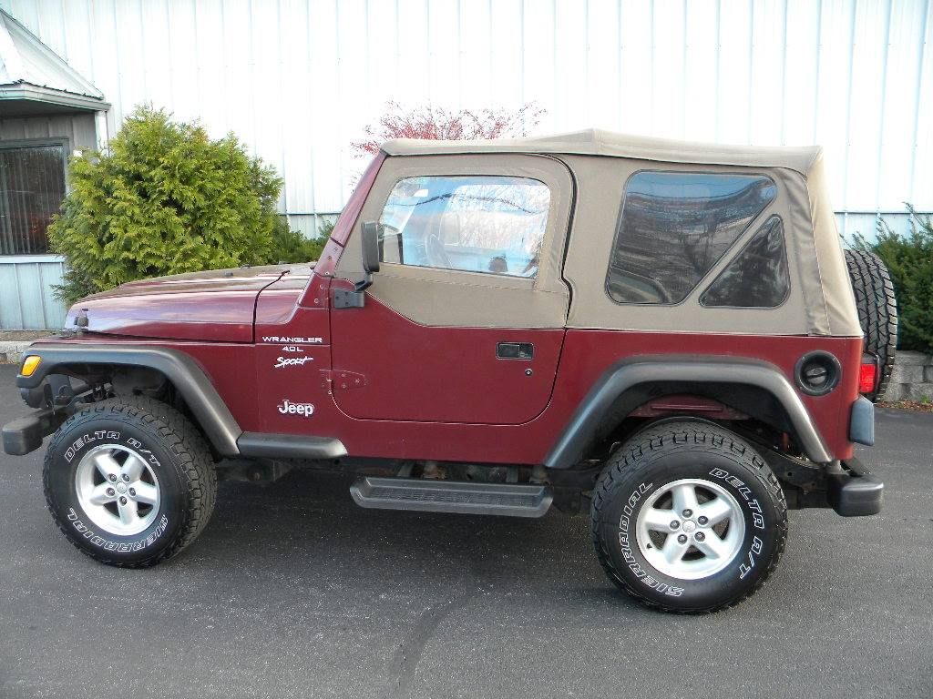 Maroon Jeep Wrangler with tan soft top and spare tire, parked outside.