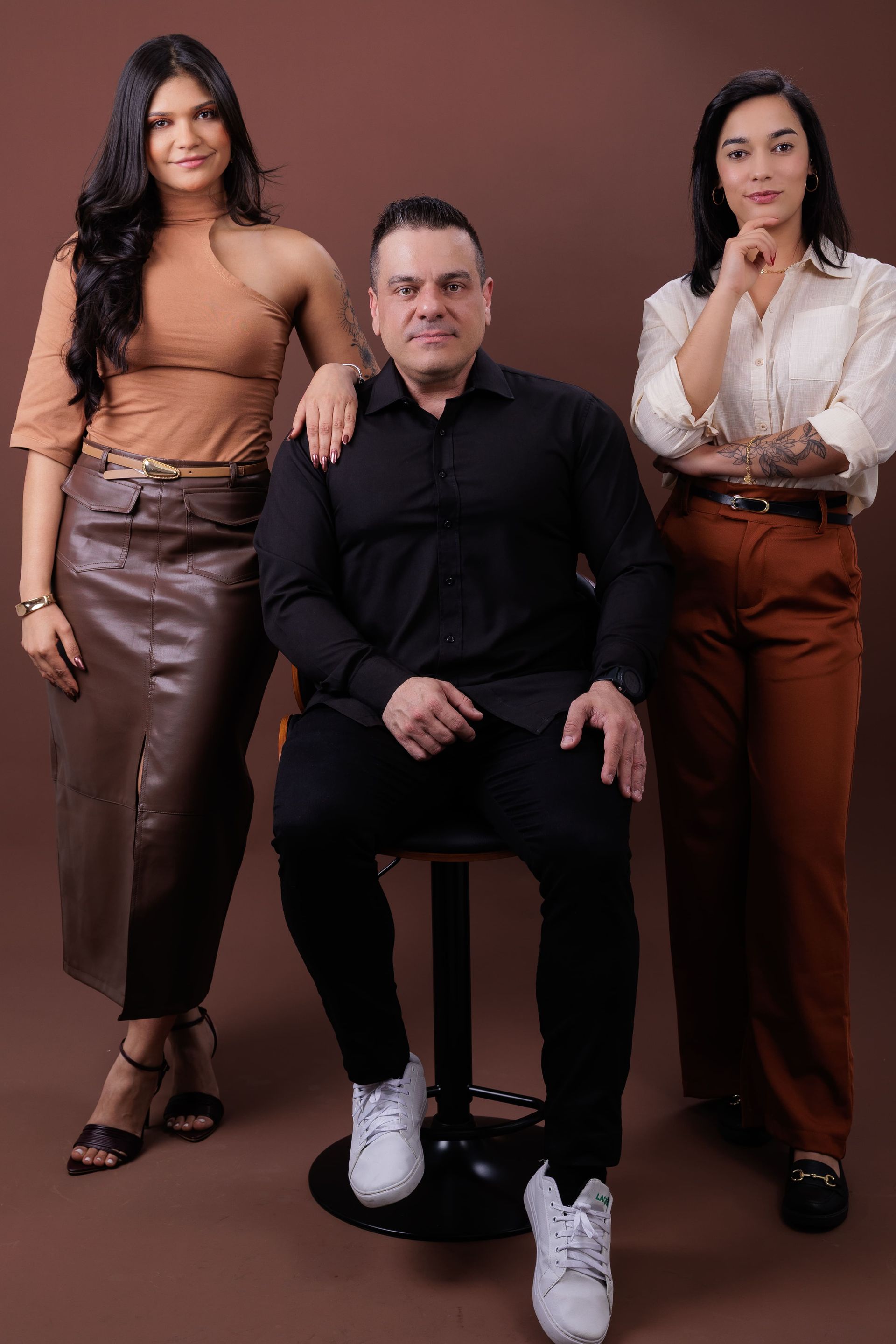 Três pessoas posando em frente a um fundo marrom. Homem sentado, ladeado por duas mulheres. Roupas marrons, pretas e brancas.