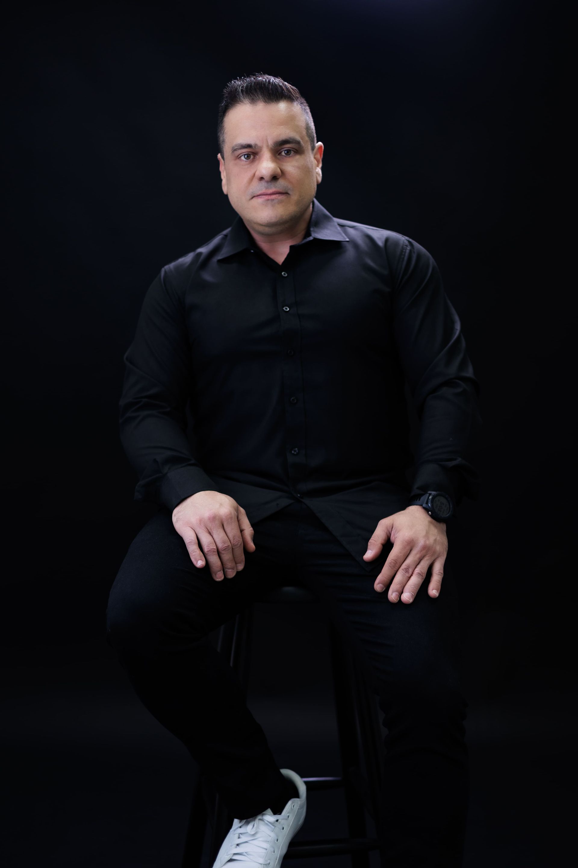 Homem de roupa preta sentado, olhando para frente, fundo escuro.