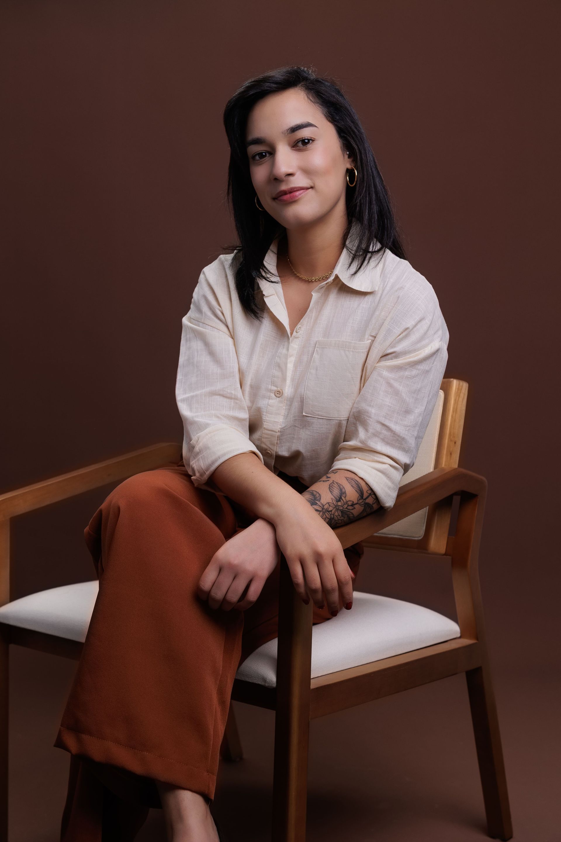 Mulher sentada em uma cadeira de madeira, vestindo camisa bege e calças cor de ferrugem