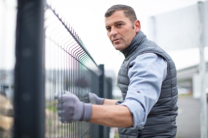 Man installing a black wire fence outdoors, wearing a vest and gloves.