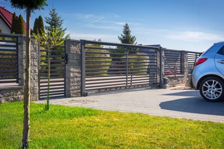 Gray car in a driveway next to a closed black metal gate and stone fence on a sunny day.