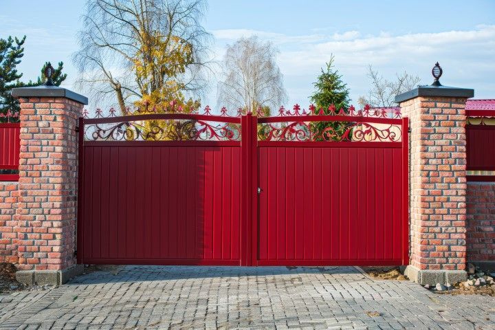 Red metal double gate between brick pillars, ornate top, cobblestone driveway.