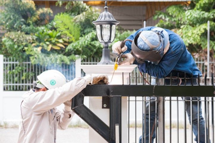 Two welders working on a black metal gate outdoors. One wears a helmet, the other a welding mask.