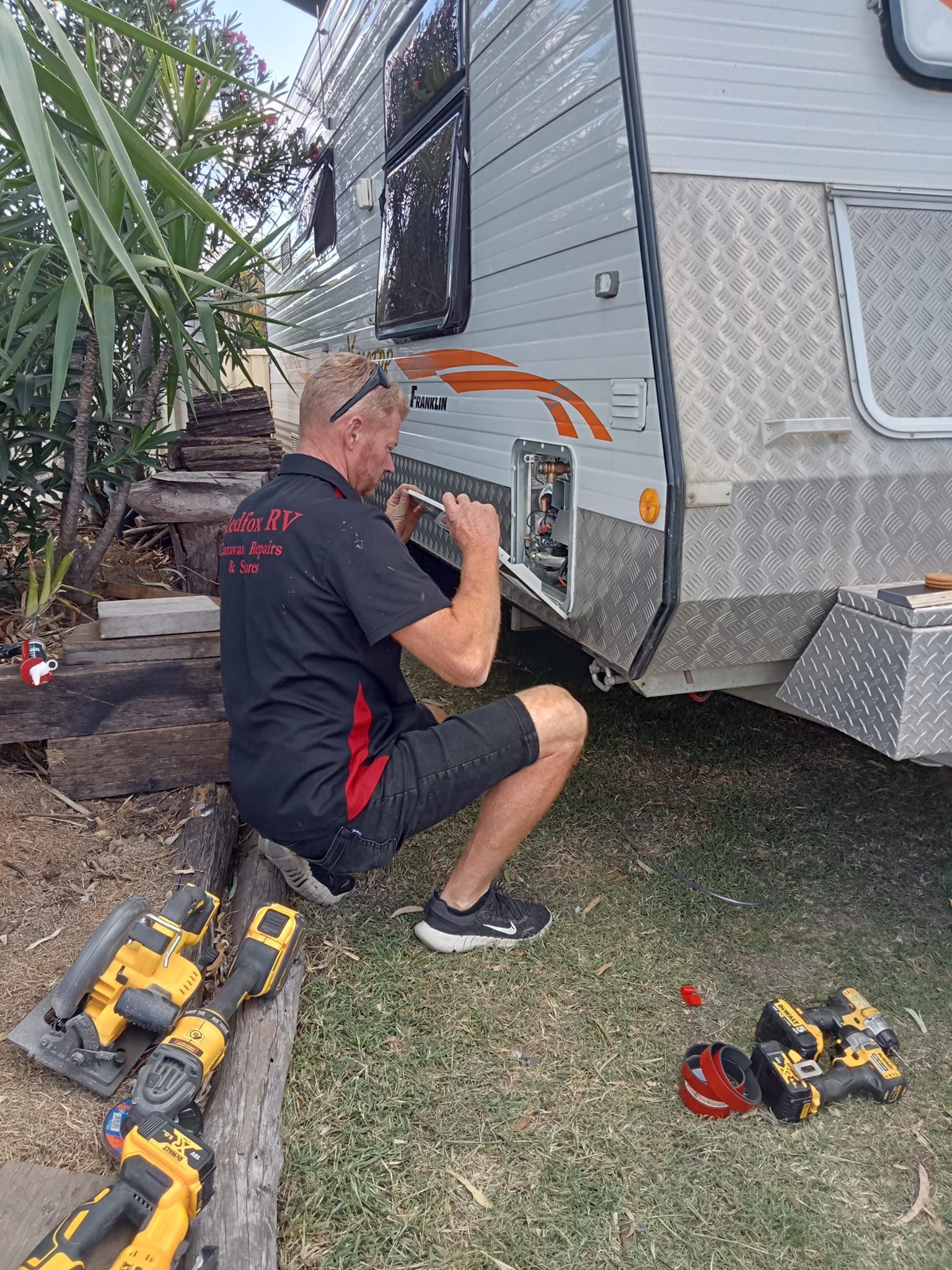 Man repairing a caravan outdoors, kneeling on grass. He is using tools.