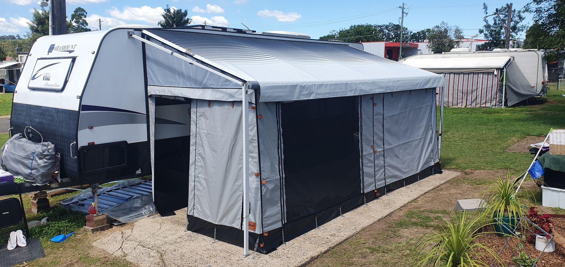 A camping setup with a camper trailer, awning, and tent-like enclosure in a grassy area under a blue sky.